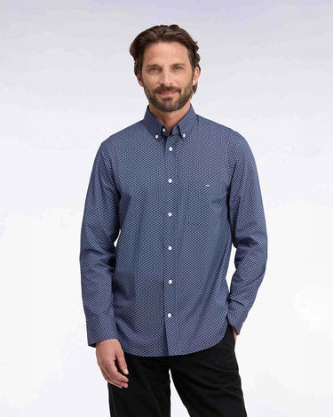 A man wearing a blue and white micro-patterned long-sleeve button-down shirt, paired with black pants, standing against a light gray studio background.