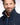 A close-up portrait of a man with a beard wearing a Eden Park Nylon Blouson Jacket, Navy over a tan collared shirt, against a light studio background.