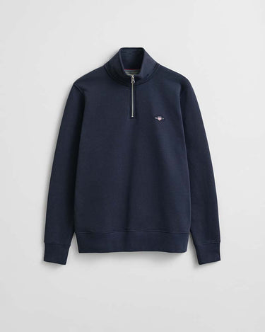 A GANT Shield Half Zip Sweat, Evening Blue with a mock neck collar and a small embroidered winged logo on the left chest, displayed against a light gray background.