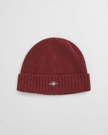 A GANT Shield Wool Beanie in Plumped Red in a deep burgundy red, featuring a small embroidered shield logo on the front of the folded cuff, set against a plain light gray background.