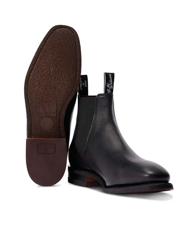 A pair of R. M. Williams Comfort Craftsman Boot Wide Fit, Black, with one boot standing upright to show the brown textured outsole and the other angled to show the side and toe, on a white background.