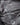 A close-up of a dark gray satin or silk-like fabric with a large metallic silver 'STRELSION' logo printed vertically, with smaller repeated logos across the surface.