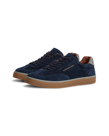 A pair of Tommy Hilfiger Luxe Suede Trainers, Navy with brown gum soles, navy laces, and gold 'TOMMY HILFIGER' branding on the side, set against a plain white background