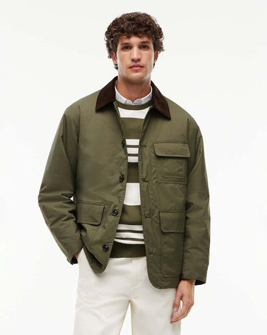 A man wearing an olive green Tommy Hilfiger Corduroy Collar Padded Ripstop Barn Jacket, layered over a striped sweater and white trousers, photographed against a plain white studio background.