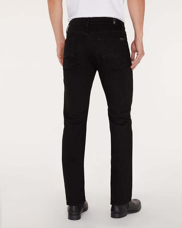 Rear view of a person wearing 7 For All Mankind Slimmy Jeans, Black with a white shirt and black shoes, photographed against a clean white studio background.