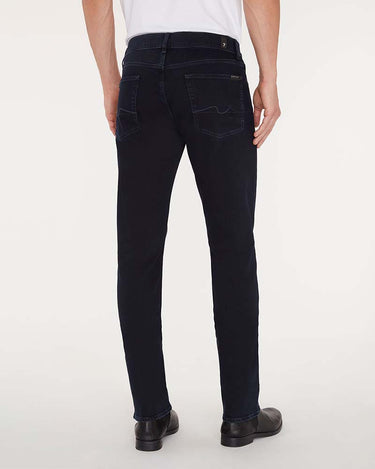 Rear view of a person wearing 7 For All Mankind Slimmy Jeans, Blue Black, a white shirt, and black shoes against a plain light background.