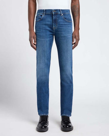 A person wearing 7 For All Mankind Slimmy Jeans, Signal Blue with a white top and black leather shoes, shown from the waist down against a light gray studio background.