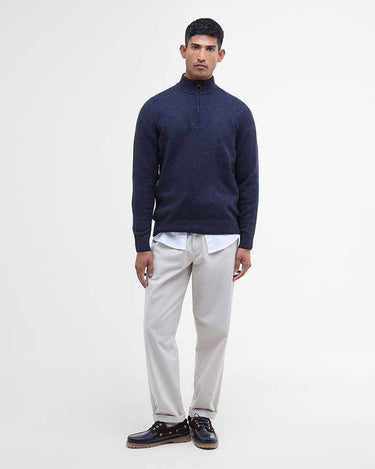 A man modeling a Barbour Tisbury Half Zip Jumper in Navy sweater, paired with off-white trousers, a white shirt, and dark leather shoes, against a plain white studio background.