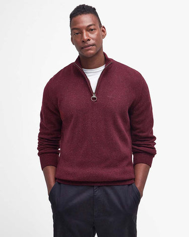 A man wearing a burgundy Barbour Tisbury Half Zip Jumper in Ruby over a white t-shirt and dark trousers, standing against a plain white studio background.