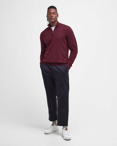 A man modeling a Barbour Tisbury Half Zip Jumper in Ruby sweater, dark charcoal trousers, and white sneakers against a plain light gray studio background.