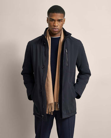 A man wearing a Bugatti All-Season Jacket, Navy over a navy sweater and a beige scarf, standing against a solid light beige background.