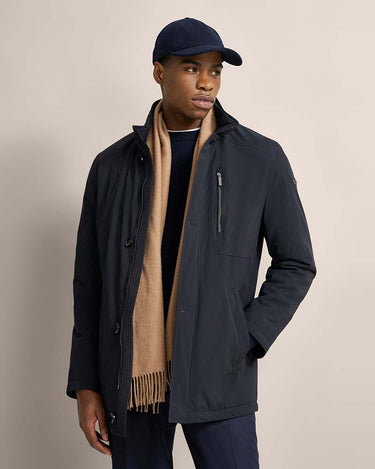 A man wearing a Bugatti All-Season Jacket, Navy, a tan scarf, a navy baseball cap, and navy trousers, standing against a solid beige studio background.
