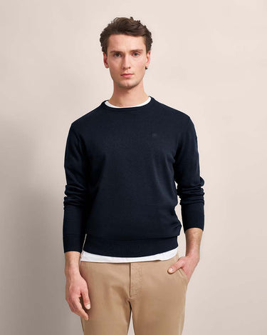 A man wearing a Bugatti Organic Cotton Crew Neck, Navy over a white undershirt and beige trousers, standing against a plain beige studio background.