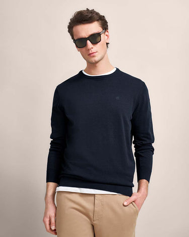 A man wearing a Bugatti Organic Cotton Crew Neck, Navy sweater with a white inner collar, styled with khaki pants and sunglasses against a solid beige background.