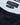 Close-up of a Bugatti Organic Cotton Crew Neck, Navy sweater showing the light blue knit body, a dark navy ribbed collar, and two black brand labels reading 'bugatti' and 'premium cotton'.