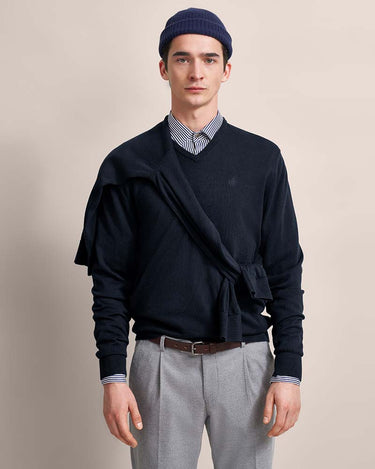 Man wearing a Bugatti V Neck Jumper, Navywith a belt, striped shirt, and gray pants on a beige background