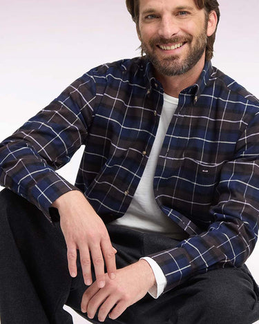 A smiling man wearing a navy blue plaid button-down shirt over a white t-shirt and dark pants, seated against a light studio background.