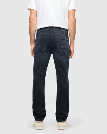 Rear view of a person wearing Mac-Jeans Arne Slim Pipe Jeans, Dark Blue a white t-shirt, and off-white sneakers against a light gray studio background.