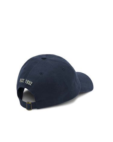A R.M. Williams Longhorn Twill Cap, Blue Wash shown from a rear three-quarter angle, featuring 'EST. 1932' embroidered in gold on the side and a gold-toned metal buckle on the back strap, set against a plain white background.