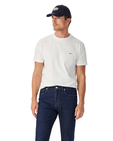 A man wearing a R.M. Williams Parson T-Shirt, White with a small embroidered bird logo on the left chest, paired with dark blue jeans and a navy blue baseball cap.