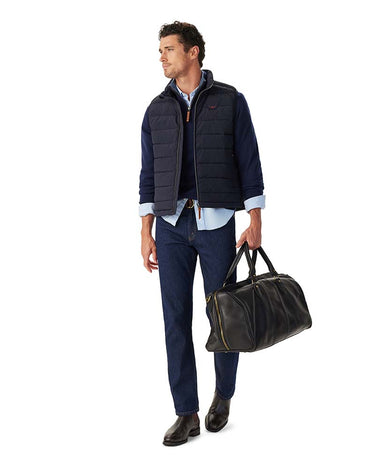 A man wearing a R.M. Williams Rydal Padded Gilet, Navyover a light blue button-down shirt, paired with dark blue jeans and black leather dress shoes, holding a black leather duffel bag with gold zippers. The photo is taken against a solid white background.