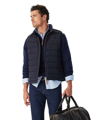 A man wearing a R.M. Williams Rydal Padded Gilet, Navy over a light blue button-down shirt and dark jeans, holding a black duffel bag against a plain white background.