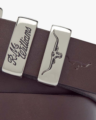 Close-up of a R.M. Williams Slim Drover Belt, Chestnut with polished silver-tone metal buckles engraved with the R.M. Williams brand name and logo.