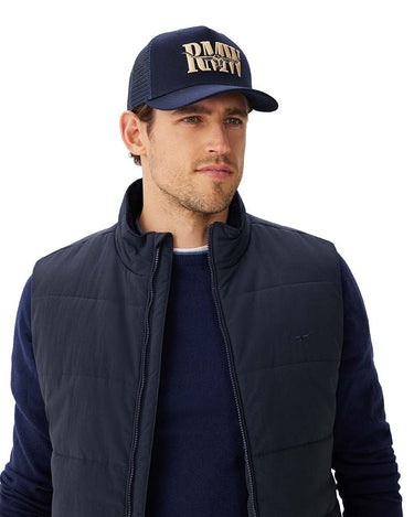 A man wearing a navy blue puffer vest over a matching navy long-sleeve shirt and a R.M. Williams Steerhead Trucker Cap, Navy with gold embroidered text, photographed against a plain white background.