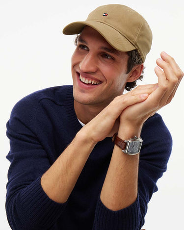 A smiling man wearing a Tommy Hilfiger 1985 Brushed Baseball Cap, Camel with a small embroidered logo, a navy blue knit sweater, and a brown leather strap watch, posed against a plain white background.