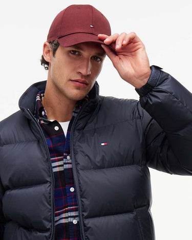 A man wearing a dark navy blue puffer jacket, a Tommy Hilfiger Flag Cotton Cap, Red Wine, and a red, blue, and white plaid shirt, posed against a clean white background. The jacket features the small embroidered Tommy Hilfiger flag logo on the left chest.