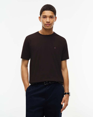 A man wearing a Tommy Hilfiger Flag Cotton T-Shirt, Brownwith a small embroidered Tommy Hilfiger flag logo on the left chest, paired with dark trousers, against a plain white studio background.