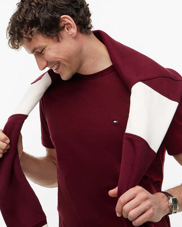 A man wearing a Tommy Hilfiger Flag Cotton T-Shirt, Wine with contrasting white color-block panels on the shoulders and sleeves. The shirt features a small embroidered Tommy Hilfiger flag logo on the left chest.