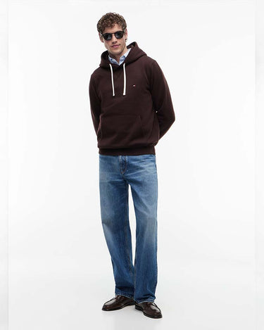 A man wearing a Tommy Hilfiger Peached Fleece Contrast Drawstring Hoodie with white drawstrings, blue jeans, and brown leather shoes, standing against a plain white studio background.